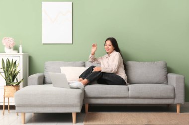 Dizüstü bilgisayarlı genç Asyalı kadın evdeki gri koltukta sohbet ediyor.