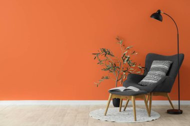 Ayak taburesi, ev bitkisi ve turuncu duvarın yanında standart lamba olan şık gri koltuk.