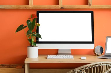 Modern bilgisayar ve turuncu duvarın yanında anthurium çiçeği olan rahat çalışma alanı, yakın plan.