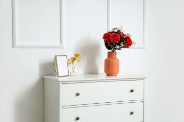 Beyaz duvarın yanındaki çekmecelerin üzerinde güzel yapay çiçekler olan vazo.