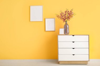 Çekmecelerin üzerinde güzel çiçekler ve turuncu duvarın yanında boş çerçeveler olan vazo.