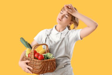 Sarı arka planda farklı olgun sebzelerle dolu sepeti olan yorgun genç bir kadın çiftçi.