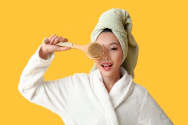 Sarı arka planda duş aldıktan sonra masaj fırçasıyla bornozlu genç Asyalı kadın.