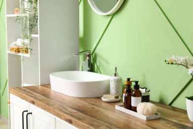 Banyonun içinde raf ünitesi, çekmeceler ve yeşil duvarın yanında lavabo var.