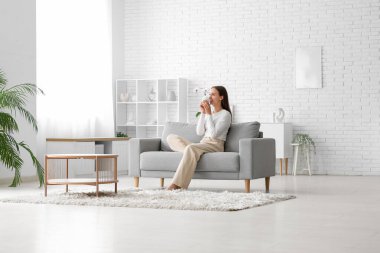 Genç ve güzel bir kadın evdeki rahat koltukta kahve içiyor.