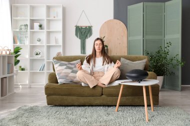 Genç bir kadın evinde glukofonla kanepede meditasyon yapıyor.