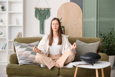 Genç bir kadın evinde glukofonla kanepede meditasyon yapıyor.