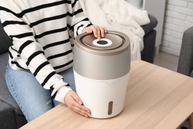 Genç bir kadın siyah koltukta oturuyor ve oturma odasındaki modern nemlendiriciyi açıyor.