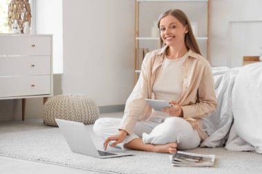 Yatak odasında dizüstü bilgisayar ve tablet kullanan genç bir kadın.