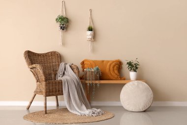 Yastıklı ahşap bank, ev bitkileri ve bej duvarın yanındaki hasır koltuk.