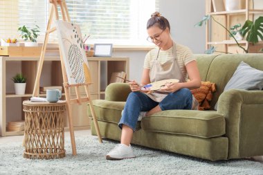 Ev stüdyosundaki kanepede resim yapan genç bir kadın sanatçı.