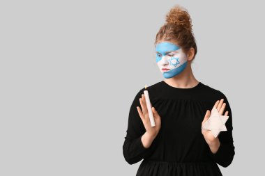 Yüzü boyalı, elinde yanan bir mum ve gri arka planda Yahudi rozeti olan genç bir kadın. Uluslararası Soykırım Anma Günü
