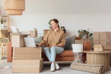 Genç bir kadın yeni evindeki koltukta dizüstü bilgisayar kullanıyor.