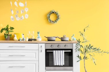 Fırın, çatal bıçak ve Paskalya süslemeleriyle dolu mutfak tezgahı sarı duvarın yanında.