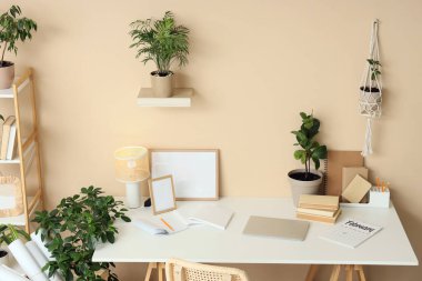 Bej duvarın yanında modern dizüstü bilgisayar, kitap ve ev bitkileriyle rahat çalışma alanı