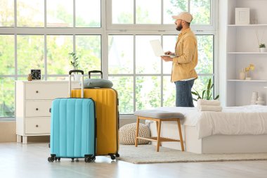 Otel odasının penceresinin yanında dizüstü bilgisayar kullanan bavullu bir erkek turist.