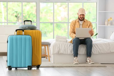 Otel odasında dizüstü bilgisayar kullanan bavullu bir erkek turist.