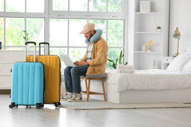 Otel odasında dizüstü bilgisayar kullanan bavullu bir erkek turist.