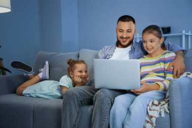 Dizüstü bilgisayarı olan mutlu bir baba ve akşam evde çizgi film izleyen kızları.