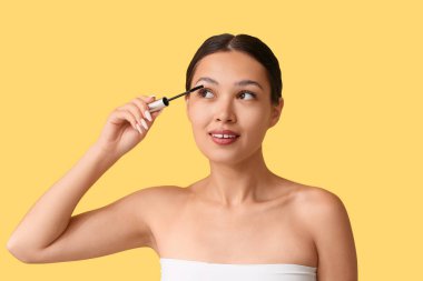 Güzel genç Asyalı kadın sarı arka plana rimel sürüyor.