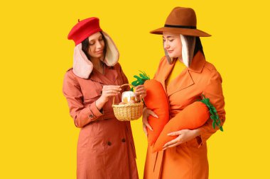 Şık paltolu ve tavşan kulaklı iki genç kadın ellerinde oyuncak havuç ve sepetle sarı arka planda Paskalya ekmeği tutuyorlar. Paskalya kutlaması