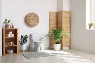 Tuvaletin içinde tuvalet, katlanan ekran, bitkiler ve raflar beyaz duvarın yanında.