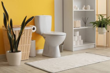 Tuvalet, çöp tenekesi, bitki ve raf ünitesi, sarı duvarın yanında.