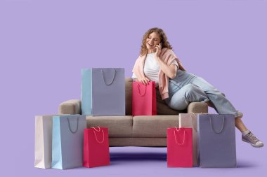 Alışveriş torbalarıyla genç mutlu bir kadın, koltukta mor arka planda telefonla konuşuyor.