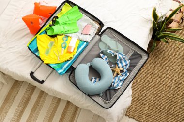 İçinde giysiler, plaj aksesuarları, pasaport, seyahat yastığı ve yatak odasında bir şişe güneş kremi olan valizi aç.