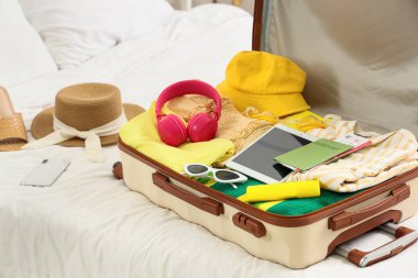 İçinde giysiler, plaj aksesuarları, pasaport, tablet, kulaklık ve yatak odasında bir şişe güneş kremi olan valizi aç.