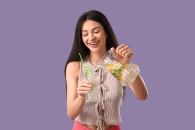 Güzel, genç, mutlu bir kadın mor arka planda turunçgil suyu dolu kavanozu dolduruyor.