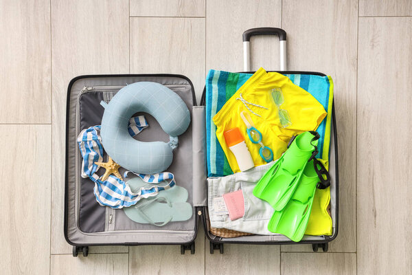 Открытый чемодан с пляжными принадлежностями, туристической подушкой, паспортом, ластами и бутылкой крема от солнца на полу в гостиной, вид сверху