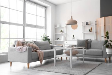 Büyük pencerenin yanında gri kanepeleri ve kahve masası olan aydınlık oturma odasının içinde.