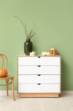 Yeşil duvarın yanında beyaz çekmeceler, balkabakları, mumlar ve ağaç dalları