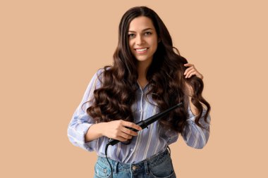 Beautiful young woman with curling iron on beige background