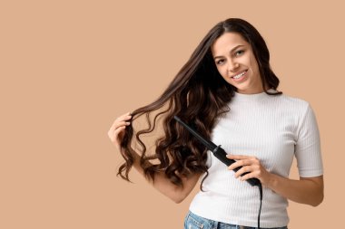 Beautiful young woman with curling iron on beige background
