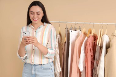 Bej arka planda giysileri olan cep telefonu kullanan genç bir kadın.