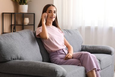 Diastemalı genç bir kadın evdeki koltukta cep telefonuyla konuşuyor.
