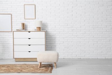 Beyaz tuğla duvarın yanında pouf, çerçeve, klozet ve hasır halı olan oturma odasının içinde.