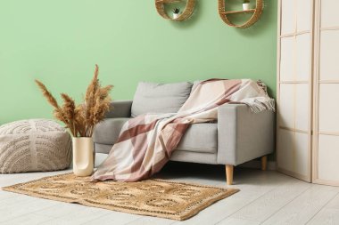 Oturma odasının içinde kanepe, katlanan perde, puf, kuru çiçekler ve yeşil duvarın yanında hasır bir halı.