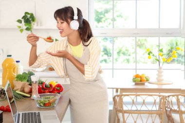 Kulaklıklı genç bir kadın mutfakta sebze salatası yiyor.