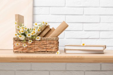 Beyaz tuğla duvarın yanındaki masada kitap ve papatya dolu sepet.