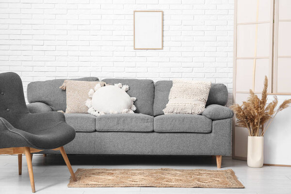 Interior of living room with sofa, armchair, dry flowers and wicker carpet on floor near white brick wall