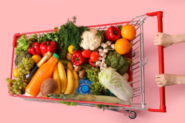 Pembe arka planda farklı yiyeceklerle dolu alışveriş arabasını iten kadın.