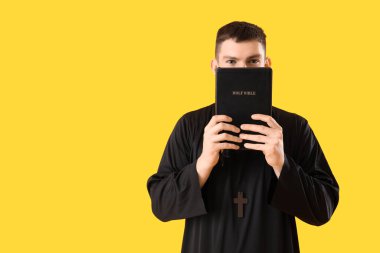 Young priest with Holy Bible on yellow background