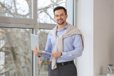 Ofisteki pencerenin yanında dizüstü bilgisayarı olan yakışıklı bir iş adamı.