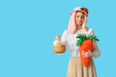 Tavşan kulaklı güzel bir kadın elinde oyuncak havuç ve sepetle mavi arka planda Paskalya ekmeği tutuyor. Paskalya kutlaması