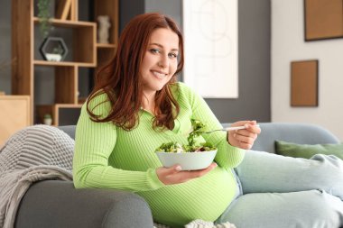 Genç hamile kadın taze sebze salatasıyla evdeki kanepede yatıyor.