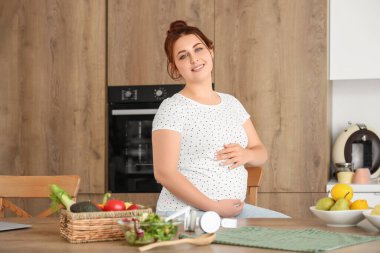 Genç hamile kadın mutfakta taze sebze salatası yiyor.