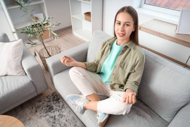 Evdeki kanepede meditasyon yapan güzel genç bir kadın.